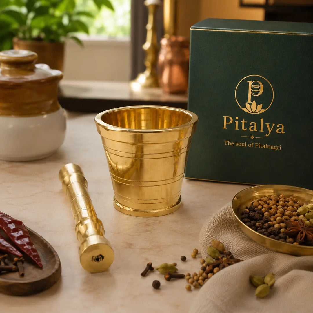 A brass mortar and pestle set with a dark green Pitalya branded box, styled on a marble surface with whole spices and dried red chilies.