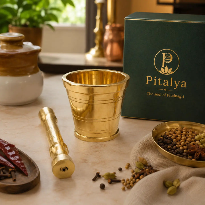 A brass mortar and pestle set with a dark green Pitalya branded box, styled on a marble surface with whole spices and dried red chilies.