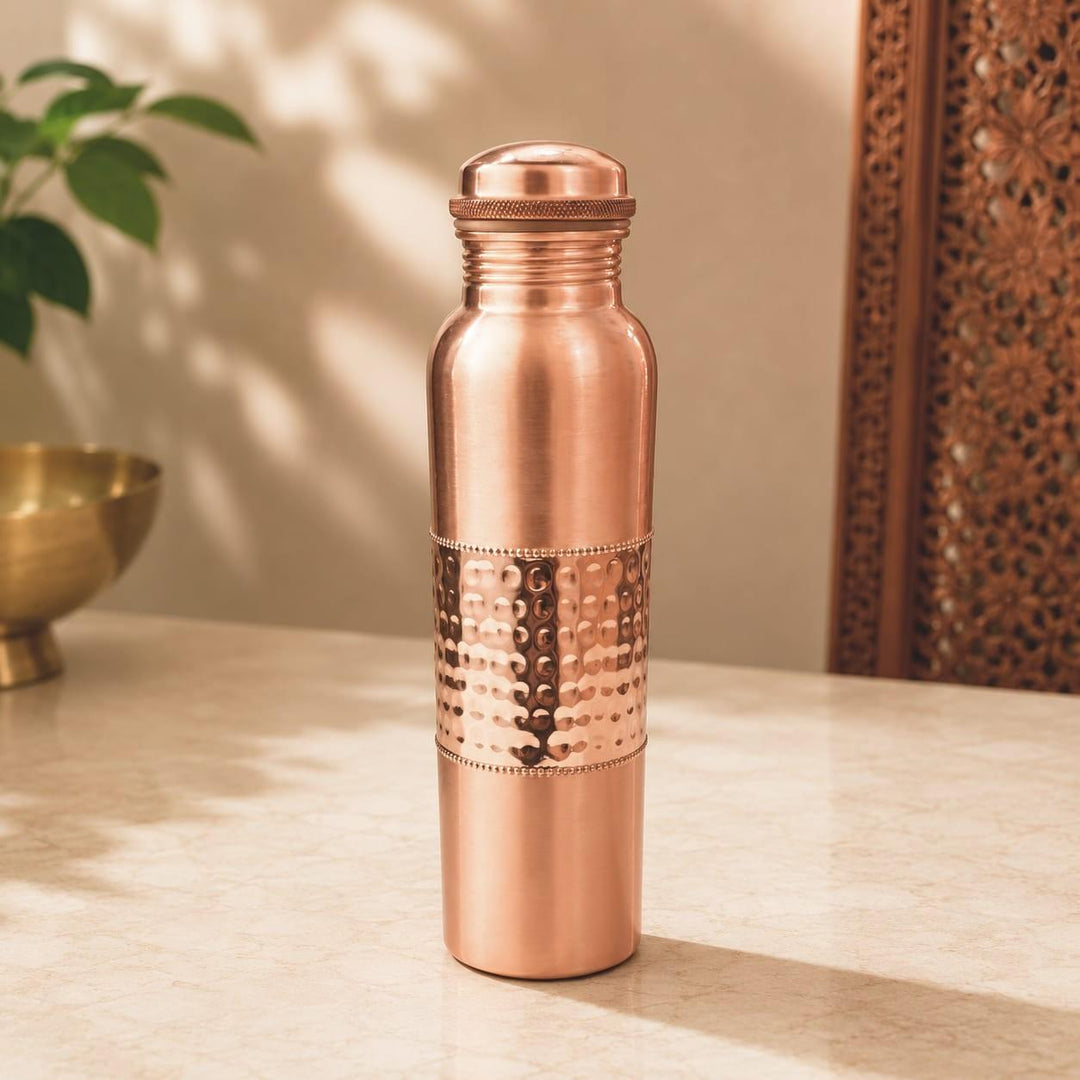 A copper water bottle with a lid, featuring a hammered metallic band around the middle, standing on a light marble surface with a warm, softly lit background.