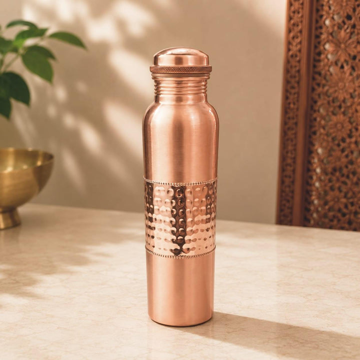 A copper water bottle with a lid, featuring a hammered metallic band around the middle, standing on a light marble surface with a warm, softly lit background.