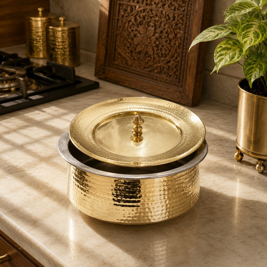 A hammered brass lidded pot with a matching brass lid and small finial, set on a marble countertop in a warm kitchen scene with decorative brass containers, a carved wooden panel, and a potted plant.