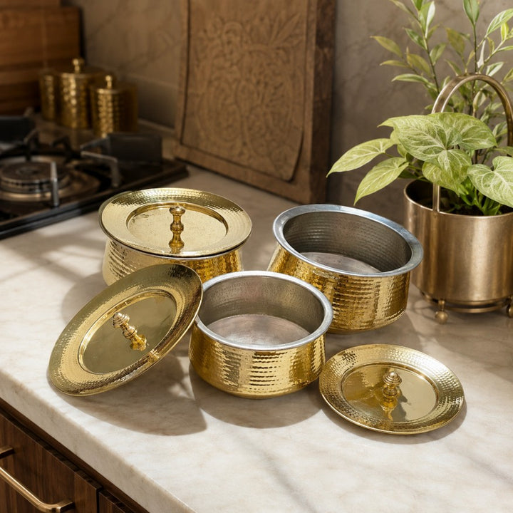 A set of gold-toned metal containers with matching lids, arranged on a marble countertop in a kitchen setting. The containers have a hammered texture and are shown in different positions, with one open container revealing a silver interior.