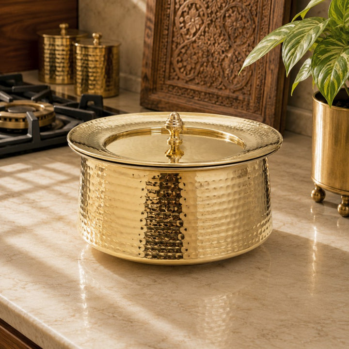 A hammered brass tawa with a matching lid, set on a marble countertop in a kitchen environment.
