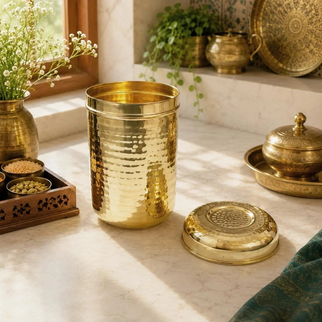 A hammered brass cylindrical container with its lid placed beside it on a marble countertop, styled in a warm, sunlit kitchen setting with other brass and bronze vessels.
