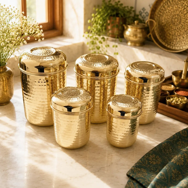 Five hammered metallic gold storage jars with matching lids arranged on a marble countertop in a warm, sunlit kitchen setting.