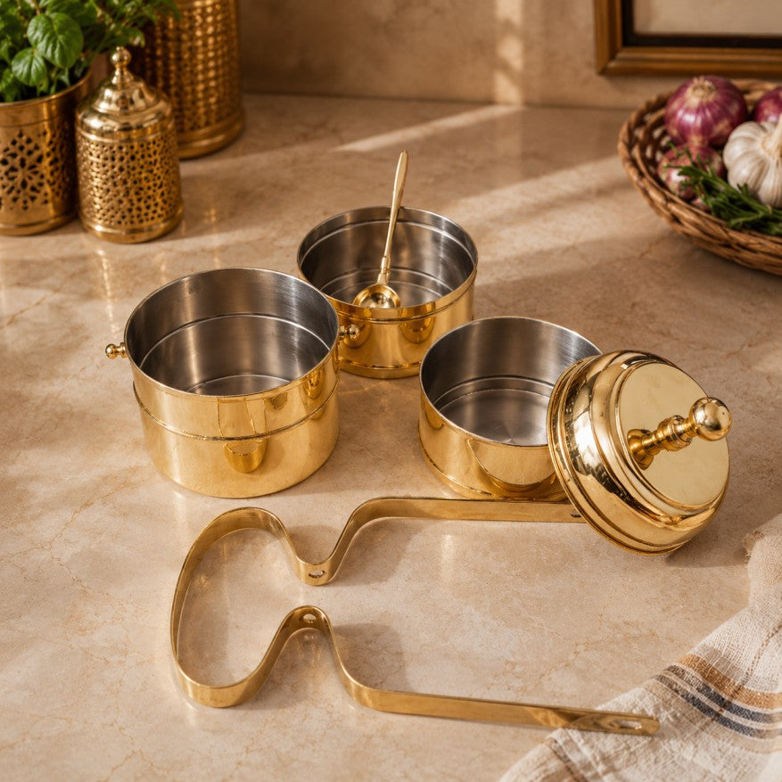 A set of brass spice containers and accessories arranged on a beige marble countertop, including two stacked spice jars, one open jar with a spoon inside, one lid with knob, and a metal scoop tool. Decorative kitchen props are visible in the background.
