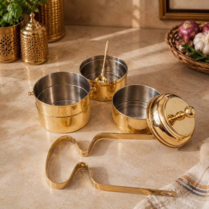 A set of brass spice containers and accessories arranged on a beige marble countertop, including two stacked spice jars, one open jar with a spoon inside, one lid with knob, and a metal scoop tool. Decorative kitchen props are visible in the background.