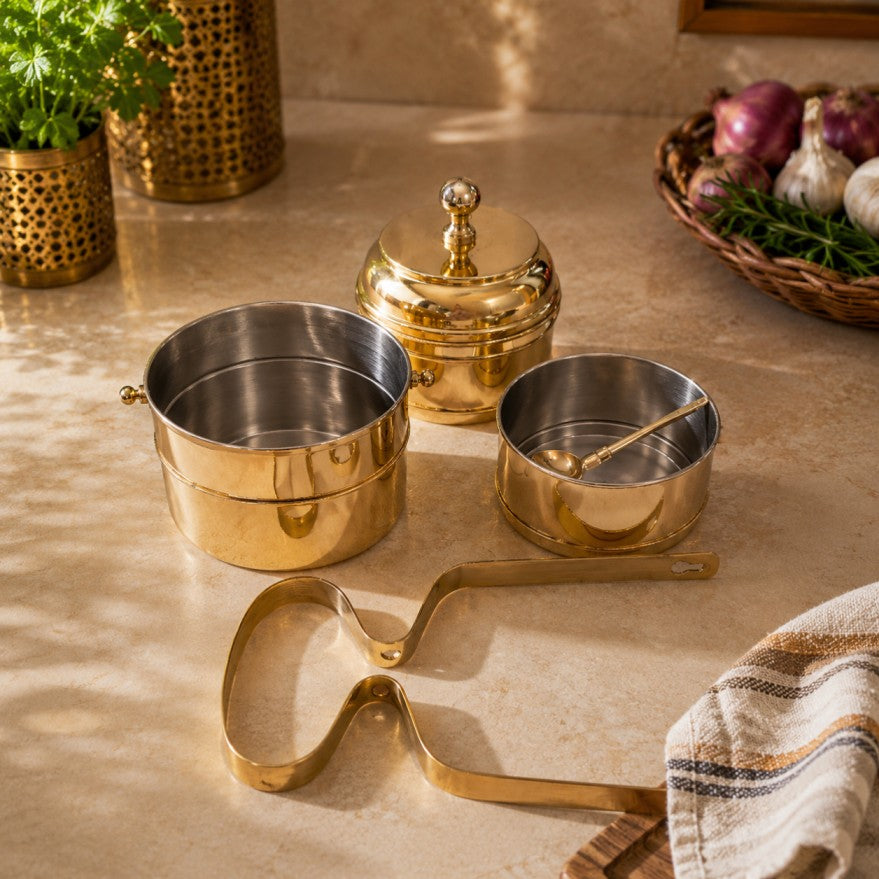 A set of polished brass and stainless steel cookware pieces arranged on a beige stone countertop, including stacked pots, a lidded pot, a small saucepan with a spoon, and a long-handled utensil, with kitchen props softly blurred in the background.