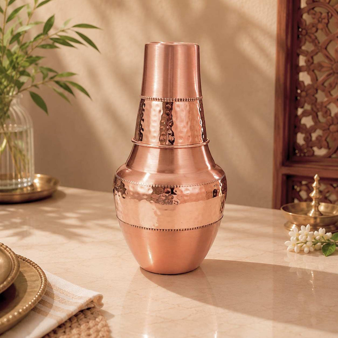 A copper-toned metal shaker or decorative vase with stacked cylindrical sections and hammered detailing, placed on a beige tabletop in a styled interior setting with plants and wooden decor.
