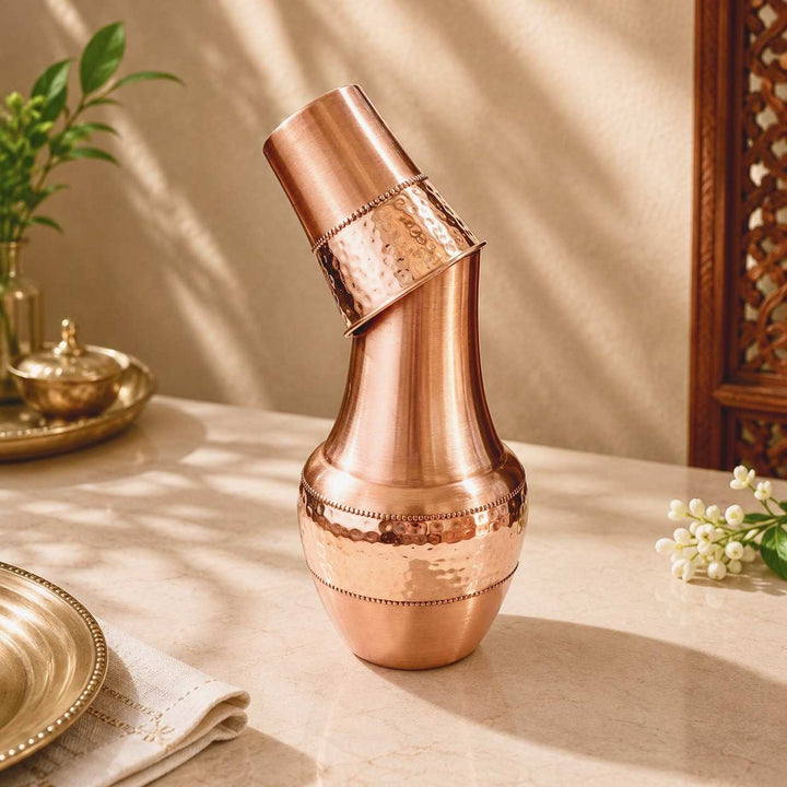 A copper-toned metal shaker bottle with a matching lid, styled on a beige tabletop with warm sunlight and shadows, surrounded by decorative props.