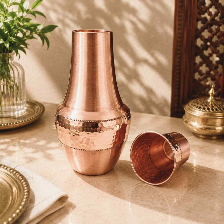A copper-toned metal cocktail shaker set with a tall neck and a separate small cup, displayed on a tabletop with warm decorative props in a real interior setting.