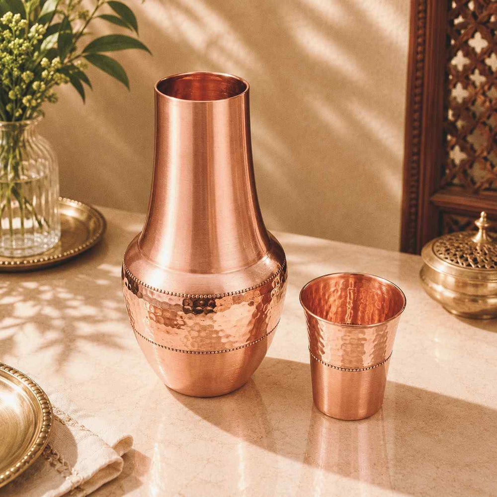A copper-toned metal decanter and matching small cup on a beige tabletop, styled with a glass vase of flowers and warm sunlight shadows.
