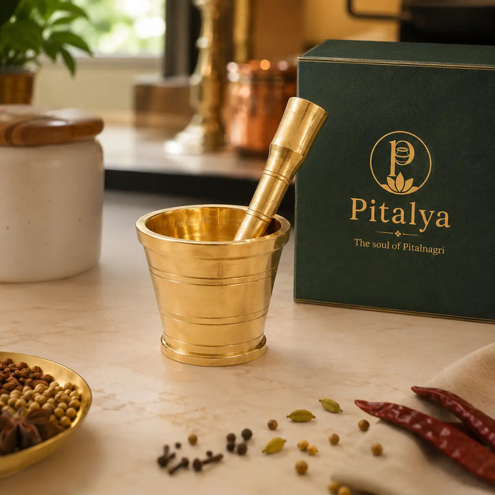 A brass mortar and pestle set displayed on a marble countertop next to its dark green Pitalya branded packaging box, with spices scattered nearby.