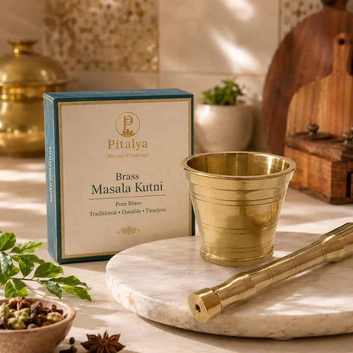 Pitalya Brass Masala Kutni set displayed on a marble tray, featuring a brass mortar and pestle alongside its teal and cream-colored product packaging box in a warm, rustic kitchen setting.