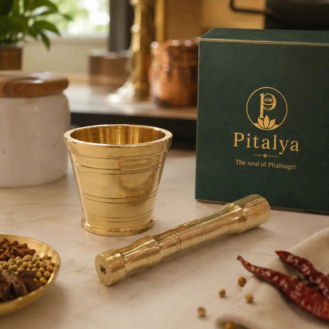 A brass mortar and pestle set displayed on a marble countertop next to its dark green Pitalya branded gift box, with spices and kitchen items softly blurred in the background.