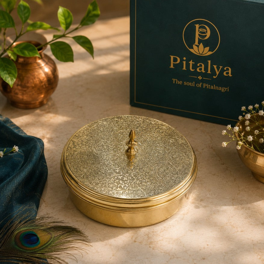 A round gold-toned lidded box with a textured metallic surface sits on a beige stone surface. Behind it is a dark teal Pitalya branded box, with decorative plants, a blue fabric cloth, and a peacock feather around it.